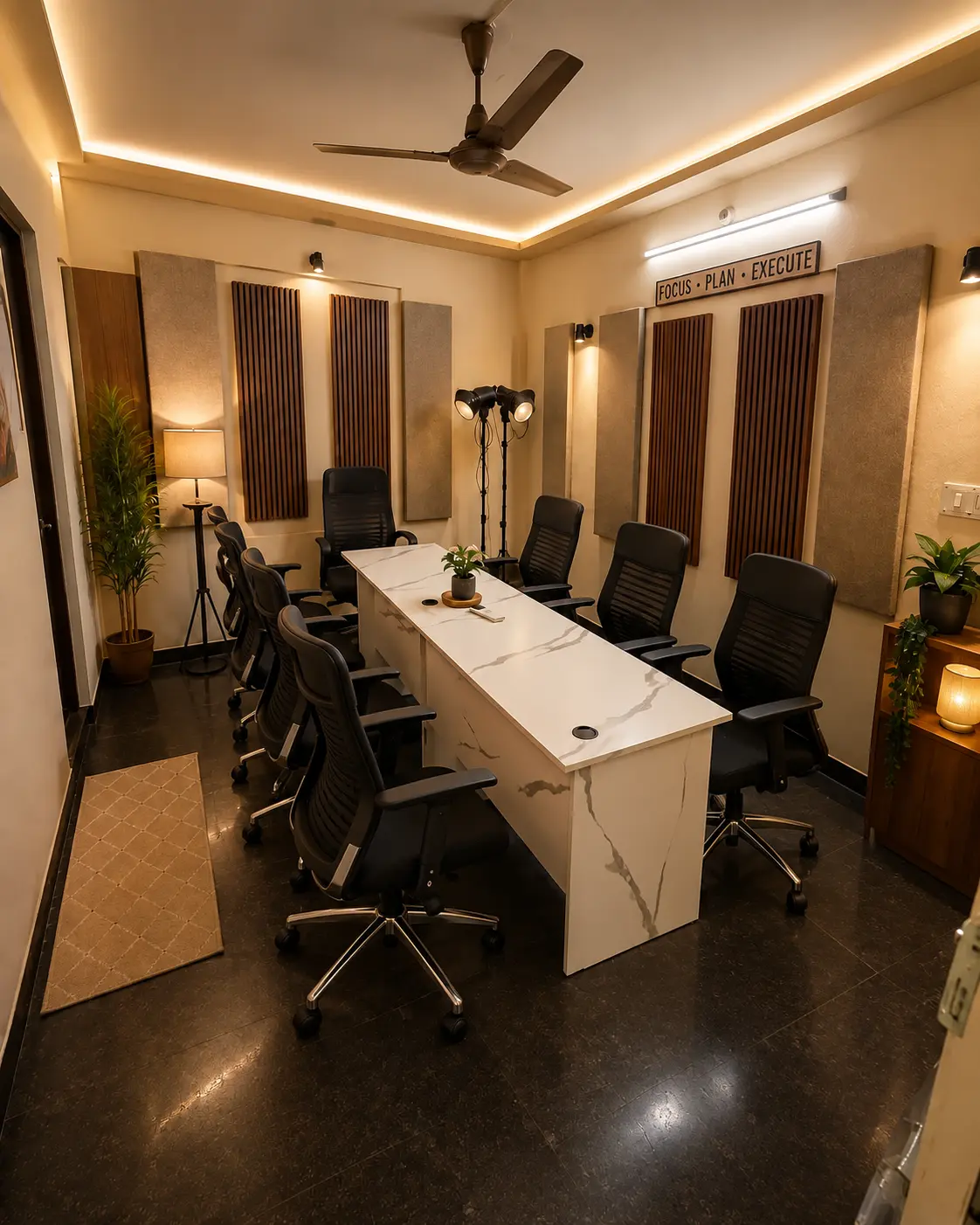A long white table down the middle of a narrow room, orange chairs along one side and black chairs along the other, acoustic panels and wood slats on the walls, two studio lights on stands at the far end, and a sign reading Focus, Plan, Execute.